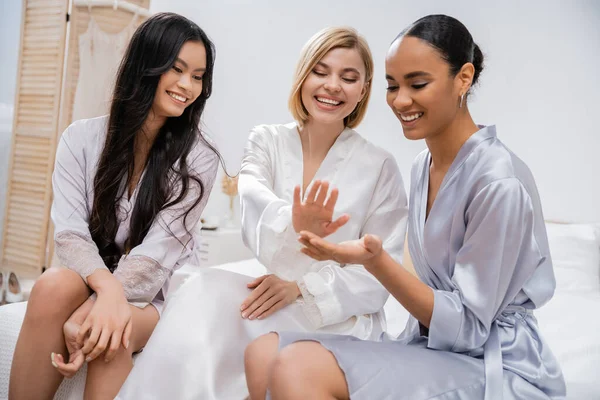 Happy blonde bride and her multicultural bridesmaids sitting on bed, showing engagement ring, happiness, bridal party, silk robes, brunette and blonde women, diversity, multicultural best friends — Stock Photo
