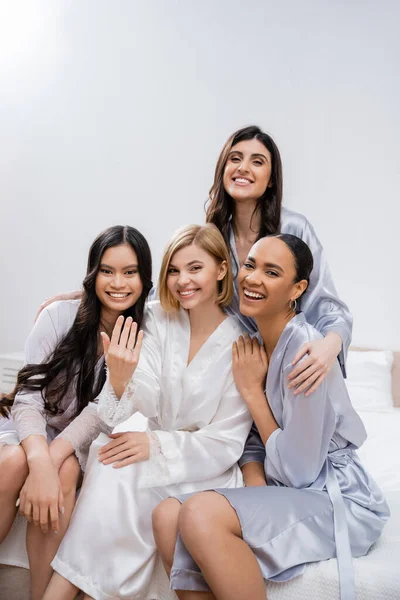 Vier Frauen, Brautparty, glückliche blonde Braut zeigt Verlobungsring in der Nähe ihrer interrassischen Brautjungfern, auf dem Bett sitzen, Glück, Seidenroben, brünett und blond, beste Freunde, Vielfalt — Stockfoto
