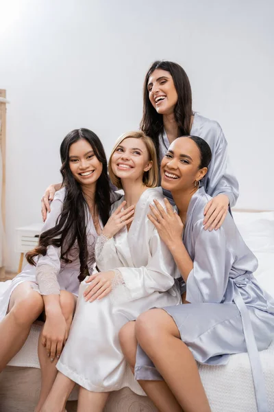 Vier Frauen, Brautparty, freudige blonde Braut und ihre gemischtrassigen Brautjungfern, die zusammen auf dem Bett sitzen, Glück, seidene Roben, Verlobungsring, brünett und blond, beste Freunde, Vielfalt — Stockfoto