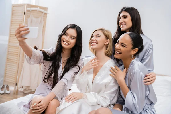 Four women, bridal party, joyful blonde bride and her interracial bridesmaids taking selfie together, happiness, silk robes, engagement ring, brunette and blonde, best friends, diversity — Stock Photo