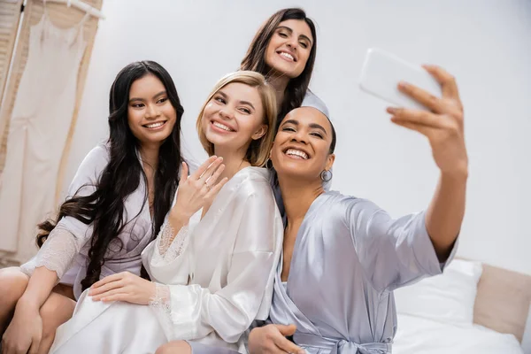 Cuatro mujeres, fiesta nupcial, novia rubia alegre y sus damas de honor interracial tomando selfie juntos, felicidad, túnicas de seda, anillo de compromiso, morena y rubia, mejores amigos, diversidad - foto de stock