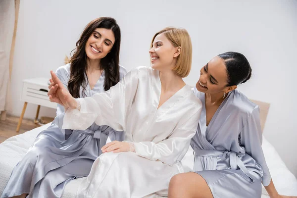 Happy blonde bride showing engagement ring to her multicultural bridesmaids, sitting on bed together, bridal shower, silk robes, brunette and blonde women, diversity, multicultural best friends — Stock Photo