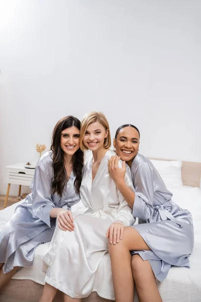 Bridal shower, happy blonde bride and her multicultural bridesmaids sitting on bed, showing engagement ring, happiness, silk robes, brunette and blonde, diversity, best friends, three women — Stock Photo