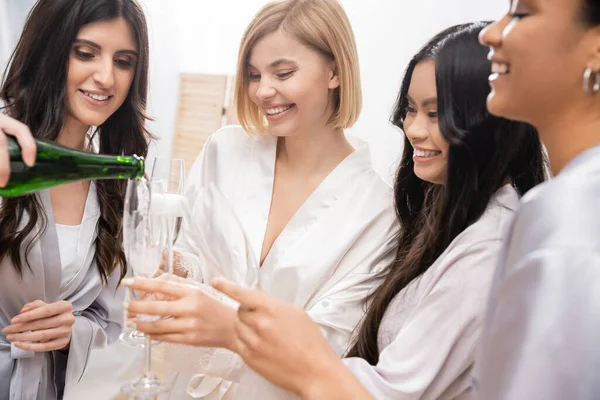 Щаслива жінка вливає шампанське в окуляри поблизу мультикультурних подруг, святкування, весела наречена і подружки нареченої, брюнетка і блондинка, різноманітність, весільний душ, кращі друзі, чотири жінки — стокове фото