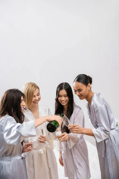 Весёлая женщина наливает шампанское в бокалы рядом с разными подругами, празднует, радостная невеста и подружки невесты, брюнетка и блондинка, разнообразие, девичник, лучшие друзья, четыре женщины, смех — стоковое фото