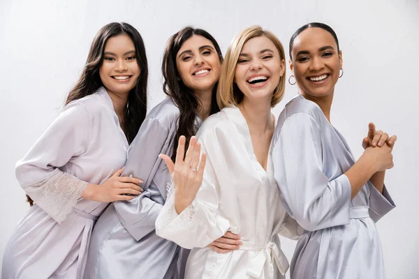 Bridal shower, four women, happy bride showing engagement ring near interracial bridesmaids in silk robes, cultural diversity, having fun together, friendship goals, brunette and blonde women — Stock Photo