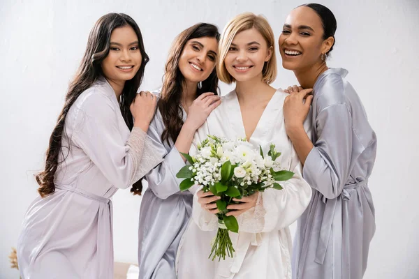 Bridal shower, four women, happy bride holding bouquet with white flowers near bridesmaids in silk robes, cultural diversity, togetherness, friendship goals, brunette and blonde women, smile and joy — Stock Photo