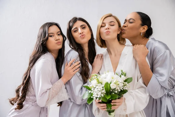Bridal shower, air kiss, four women, bride holding bouquet with white flowers near bridesmaids in silk robes, cultural diversity, togetherness, friendship goals, brunette and blonde women — Stock Photo