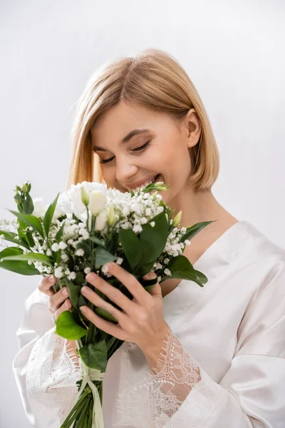 White flowers, happiness, cheerful bride with blonde hair standing in white silk robe and smelling bridal bouquet, young woman, beautiful, excitement, feminine, blissful, portrait, grey background — Stock Photo