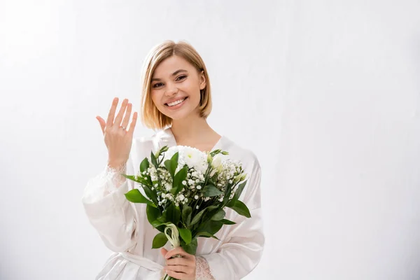 Happiness, cheerful bride with blonde hair standing in white silk robe and holding bridal bouquet, showing engagement ring, young woman, beautiful, excitement, feminine, blissful, white flowers — Stock Photo