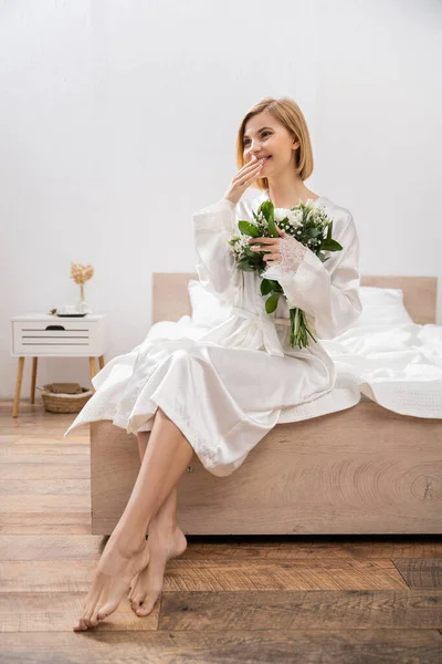 White flowers, happiness, cheerful bride with blonde hair sitting on bed and holding bridal bouquet, young woman in white robe, beautiful, excitement, feminine, wedding preparation, hand near lips — Stock Photo