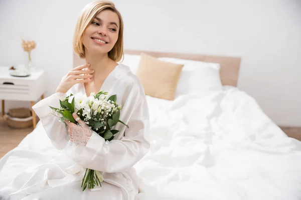 Предвкушение и счастье, веселая невеста с светлыми волосами, сидящая на кровати и держащая свадебный букет, молодая женщина в белом халате, красивая, волнительная, женственная, блаженная, свадебная подготовка — стоковое фото