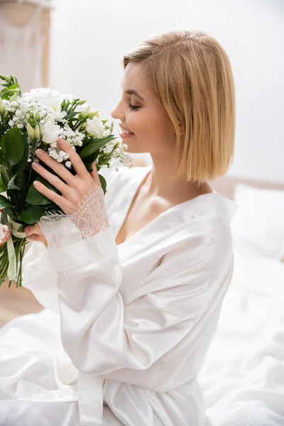 Щастя, весела наречена з світлим волоссям сидить на ліжку і пахне білими квітами, весільний букет, молода жінка в білому халаті, красива, хвилююча, жіночна, блаженна, весільна підготовка — стокове фото