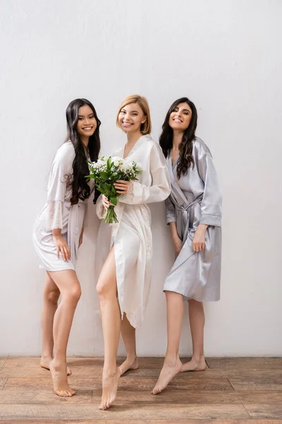 Cheerful bride with white flowers, diverse bridesmaids, bridal bouquet, cultural diversity, friendship goals, brunette and blonde women, bridal shower, smile and joy, full length — Stock Photo