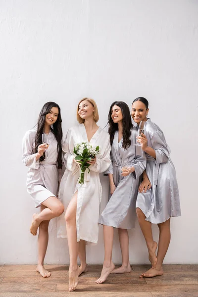 Bridal party, interracial girlfriends holding glasses with champagne, bride with white flowers, brunette and blonde women, bridesmaids, cultural diversity, positivity, bridal bouquet, grey background — Stock Photo
