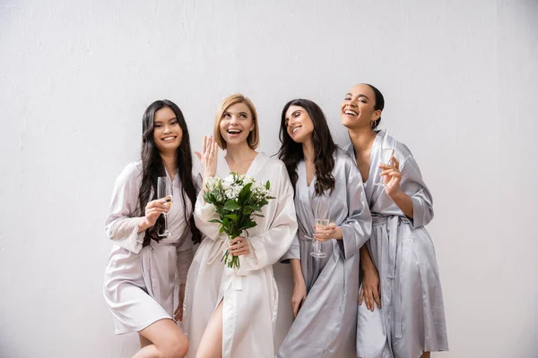Bridal party, multicultural women holding glasses with champagne, bride with white flowers showing her engagement ring, bridesmaids, diversity, positivity, bridal bouquet, grey background — Stock Photo