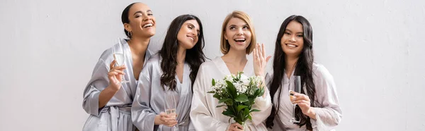 Bridal party, multicultural women holding glasses with champagne, bride with white flowers showing her engagement ring, bridesmaids, diversity, positivity, bridal bouquet, grey background, banner — Stock Photo