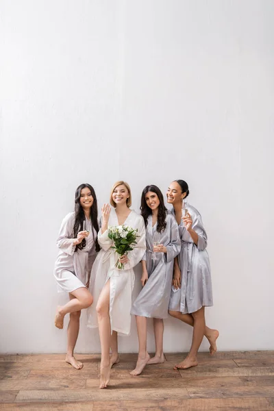 Bridal shower, multicultural women holding glasses with champagne, bride with bouquet showing her engagement ring, bridesmaids, diversity, positivity, white flowers, grey background — Stock Photo