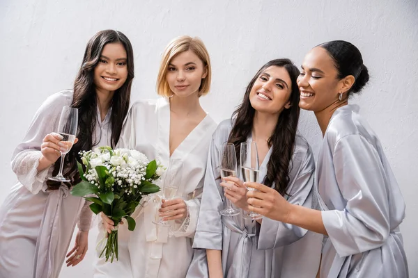 Весільний душ, щасливі мультикультурні подружки тримають окуляри з шампанським, наречена з білими квітами, брюнетка і блондинка жінки, подружки нареченої, різноманітність, весільний букет, сірий фон — стокове фото