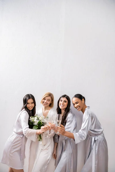 Happy multicultural girlfriends clinking glasses with champagne, bride with white flowers, brunette and blonde women, bridesmaids, diversity, positivity, bridal bouquet, grey background — Stock Photo