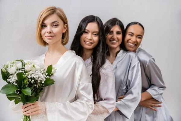 Positivity, bride and bridesmaids, happy woman holding bouquet of flowers and standing near cheerful interracial friends on grey background, racial diversity, silk robes, fashion — Stock Photo