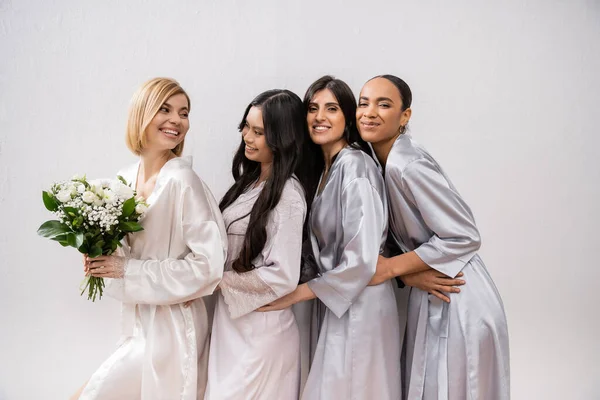 Excitement, joyful bride holding bouquet of flowers and standing near cheerful interracial bridesmaids on grey background, racial diversity, silk robes, fashion, brunette and blonde women — Stock Photo