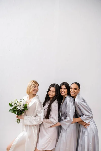 Positivity, happy bride holding bouquet of flowers and standing near cheerful interracial bridesmaids on grey background, racial diversity, silk robes, fashion, friendship, brunette and blonde women — Stock Photo
