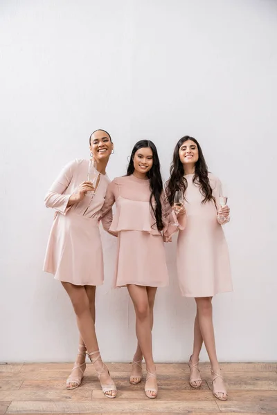 Trois demoiselles d'honneur multiculturelles, jolies femmes en robes rose pastel tenant des verres de champagne sur fond gris, diversité culturelle, mode, célébration, acclamations — Photo de stock