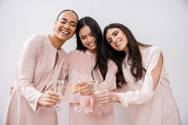 Tres damas de honor multiculturales, mujeres bonitas en vestidos de color rosa pastel tintineando copas de champán sobre fondo gris, diversidad cultural, moda, celebración, hurras - foto de stock
