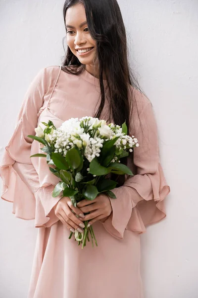 Счастливая подружка невесты в пастельно-розовом платье с букетом, азиатка с брюнетками на сером фоне, белые цветы, особый случай, свадьба, мода, улыбка и радость — стоковое фото