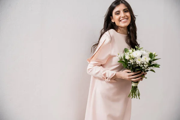 Веселая подружка невесты в пастельно-розовом платье, держа букет невесты, брюнетка на сером фоне, белые цветы, особый случай, свадьба, мода, улыбка и радость, глядя в камеру — стоковое фото