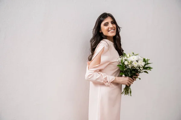 Positive Brautjungfer in pastellrosa Kleid hält Brautstrauß, brünette Frau auf grauem Hintergrund, weiße Blumen, besonderen Anlass, Hochzeit, Mode, Lächeln und Freude, Blick in die Kamera — Stockfoto