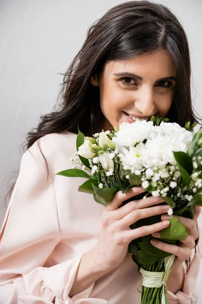 Особый случай, счастливая подружка невесты в пастельно-розовом платье держа букет, брюнетка на сером фоне, белые цветы, свадьба, мода, улыбка и радость, глядя в камеру — стоковое фото