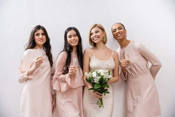 Позитивность, счастливая невеста в свадебном платье держа букет невесты и стоя рядом межрасовые подружки невесты на сером фоне, бокалы шампанского, расовое разнообразие, мода, брюнетки и блондинки — стоковое фото