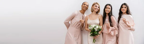 Positivity, happy bride in wedding dress holding bridal bouquet and standing near interracial bridesmaids on grey background, champagne glasses, racial diversity, fashion, banner — Stock Photo