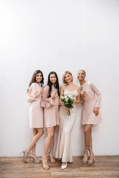Positivity, blonde bride in wedding dress holding bouquet and standing near interracial bridesmaids with champagne glasses on grey background, racial diversity, fashion, multicultural young women — Stock Photo