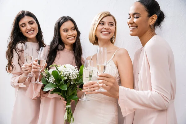 Позитивність, блондинка наречена у весільній сукні тримає букет, стоїть з міжрасовими подружками, келихами шампанського, сірим фоном, расовим розмаїттям, модою, мультикультурними молодими жінками — стокове фото