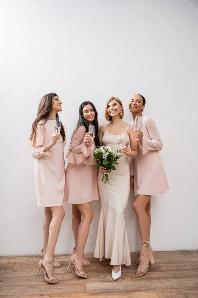 Joy, blonde bride in wedding dress holding bouquet, standing with interracial bridesmaids, champagne glasses, grey background, racial diversity, fashion, multicultural young women — Stock Photo