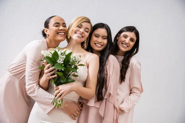 Позитивность, жизнерадостные межрасовые подружки невесты, обнимающие счастливую невесту в свадебном платье, свадебный букет, серый фон, расовое разнообразие, мода, брюнетки и блондинки, белые цветы — стоковое фото