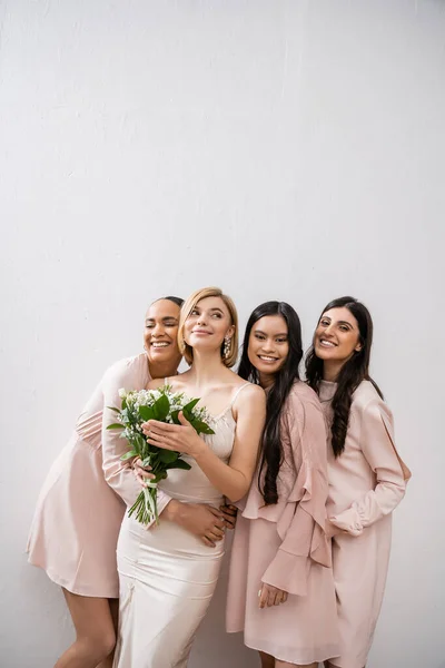 Позитивность, счастливые межрасовые подружки невесты, обнимающие невесту в свадебном платье, свадебный букет, серый фон, расовое разнообразие, мода, брюнетки и блондинки, белые цветы — стоковое фото