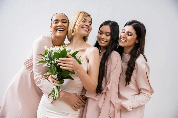 Aufregung, multikulturelle Brautjungfern umarmen glückliche Braut im Brautkleid mit Brautstrauß, grauer Hintergrund, Rassenvielfalt, Mode, brünett und blond, Frauengruppe, weiße Blumen — Stockfoto