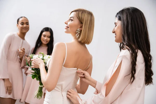 Подготовка к свадьбе, подружка невесты на молнии свадебное платье блондинки невесты, межрасовые женщины на размытом сером фоне, расовое разнообразие, мода, брюнетки и блондинки женщины, свадебный букет — стоковое фото
