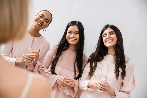 Special occasion, wedding preparations, happy multicultural bridesmaids with glasses of champagne looking at bride on grey background, admire her style, fitting, bridesmaid gowns, diversity, blurred — Stock Photo