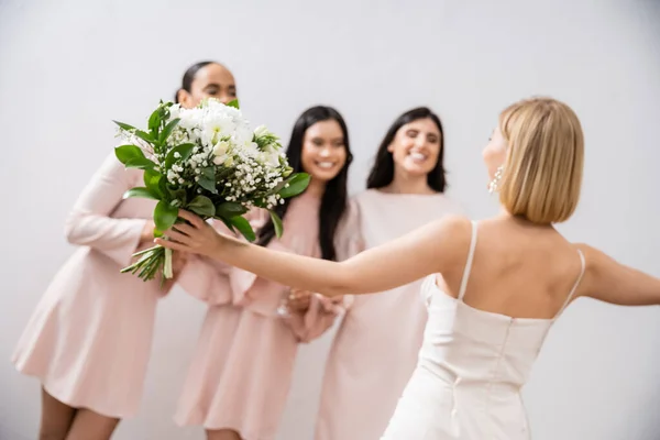 Hochzeitsvorbereitungen, fröhliche Braut mit Strauß, die neben verschwommenen multikulturellen Brautjungfern auf grauem Hintergrund steht, Kleideranprobe, Brautjungfernkleider, Hochzeitskleid, Vielfalt — Stockfoto