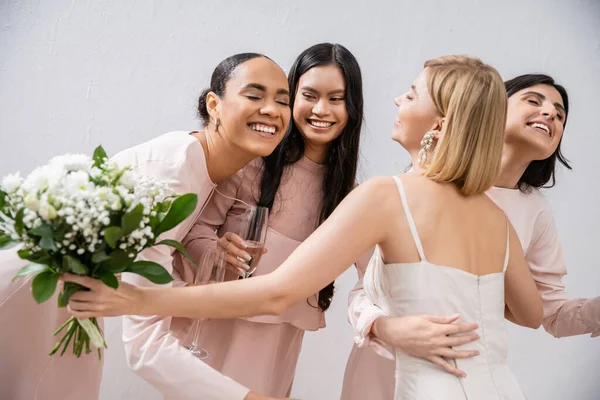 Wedding preparations, happy bride with bouquet hugging cheerful multicultural bridesmaids with champagne on grey background, dress fitting, bridesmaid gowns, wedding dress, diversity — Stock Photo