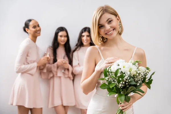 Приваблива наречена у весільній сукні, що тримає букет, стоїть біля розмитих міжрасових подружок наречених на сірому фоні, щастя, особливого випадку, блондинки та брюнетки — стокове фото