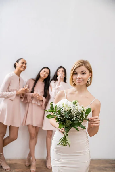 Красива наречена у весільній сукні, що тримає весільний букет, стоїть біля розмитих міжрасових подружок наречених на сірому фоні, щастя, особливого випадку, блондинки та брюнетки — стокове фото