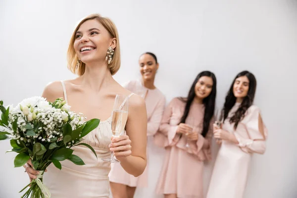 Веселая невеста в свадебном платье, держа в руках свадебный букет и бокал шампанского, стоя рядом с размытыми межрасовыми подружками невесты на сером фоне, счастье, особый случай, блондинка и брюнетка женщины — стоковое фото