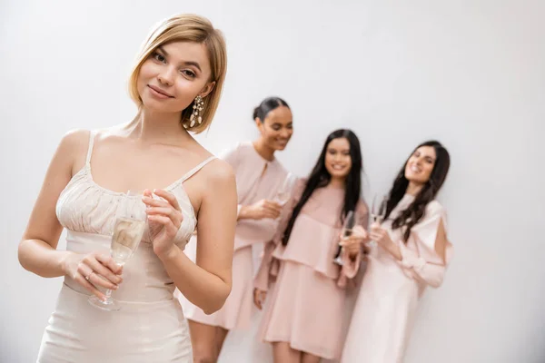 Весёлая невеста в свадебном платье с бокалом шампанского, стоящая рядом с размытыми межрасовыми подружками невесты на сером фоне, счастье, особый случай, блондинка и брюнетка — стоковое фото