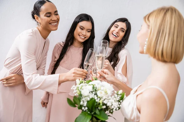 Glückliche multikulturelle Frauen, die Gläser mit Champagner klappern, Braut mit weißen Blumen, brünette und blonde Frauen, Brautjungfern, Vielfalt, Positivität, Brautstrauß, grauer Hintergrund — Stockfoto